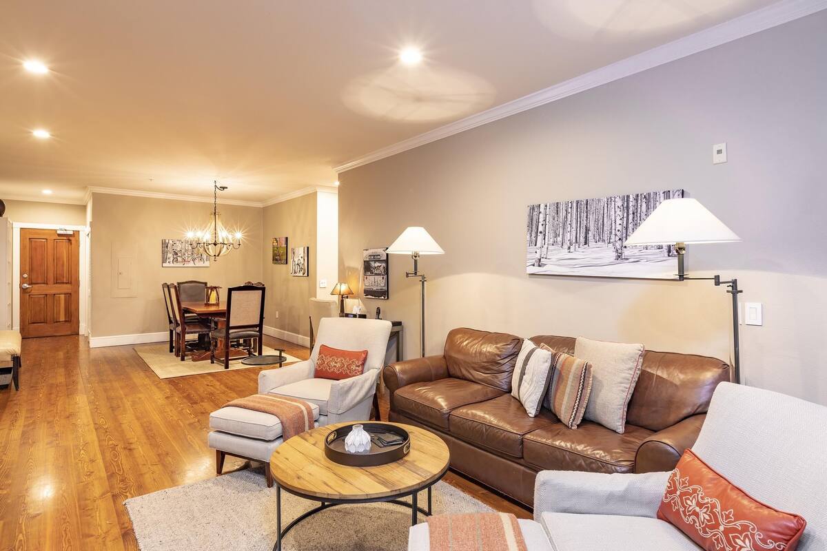 A living room with a brown leather sofa, a grey armchair, a wooden coffee table, and a dining area with a wooden table and chairs.
