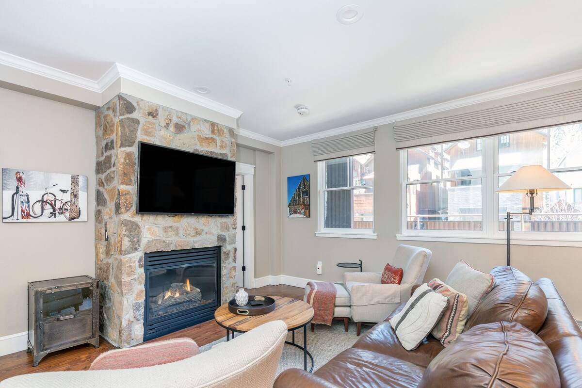 A living room with a stone fireplace, a flat-screen TV, a leather couch, and a window with blinds.