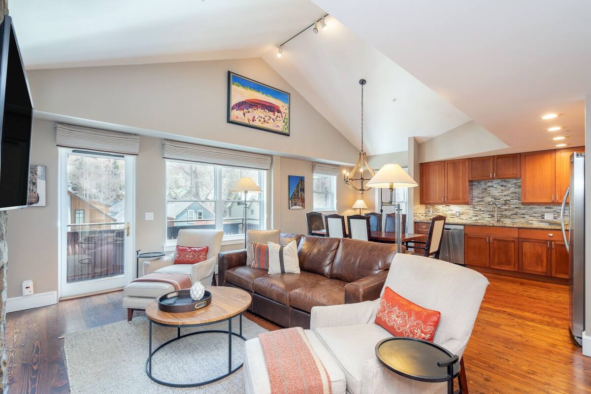 A spacious living room with a brown leather sofa, white armchairs, and a coffee table. A kitchen with wooden cabinets and a stone backsplash is visible in the background.