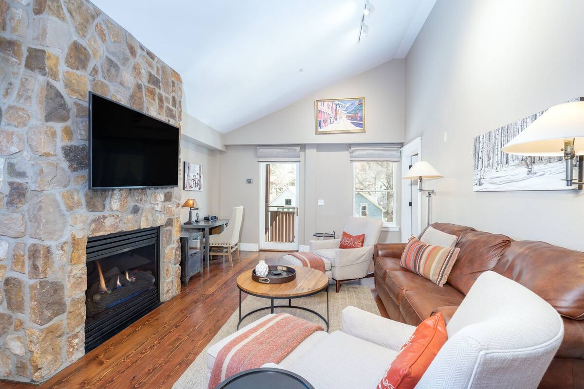 A cozy living room with a stone fireplace, a flat-screen TV, a leather sofa, and a dining area with a table and chairs.