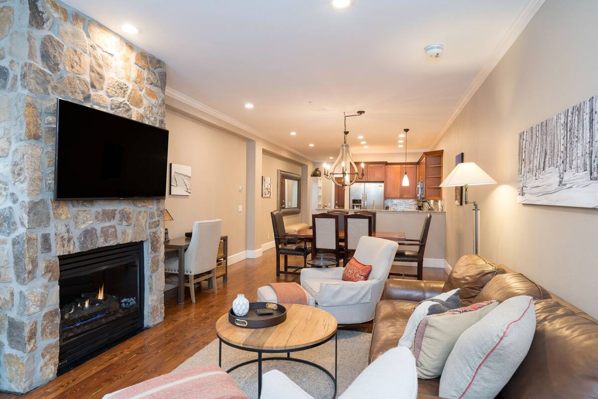 A living room with a stone fireplace, a flat-screen TV, a leather sofa, and a dining area with a chandelier.