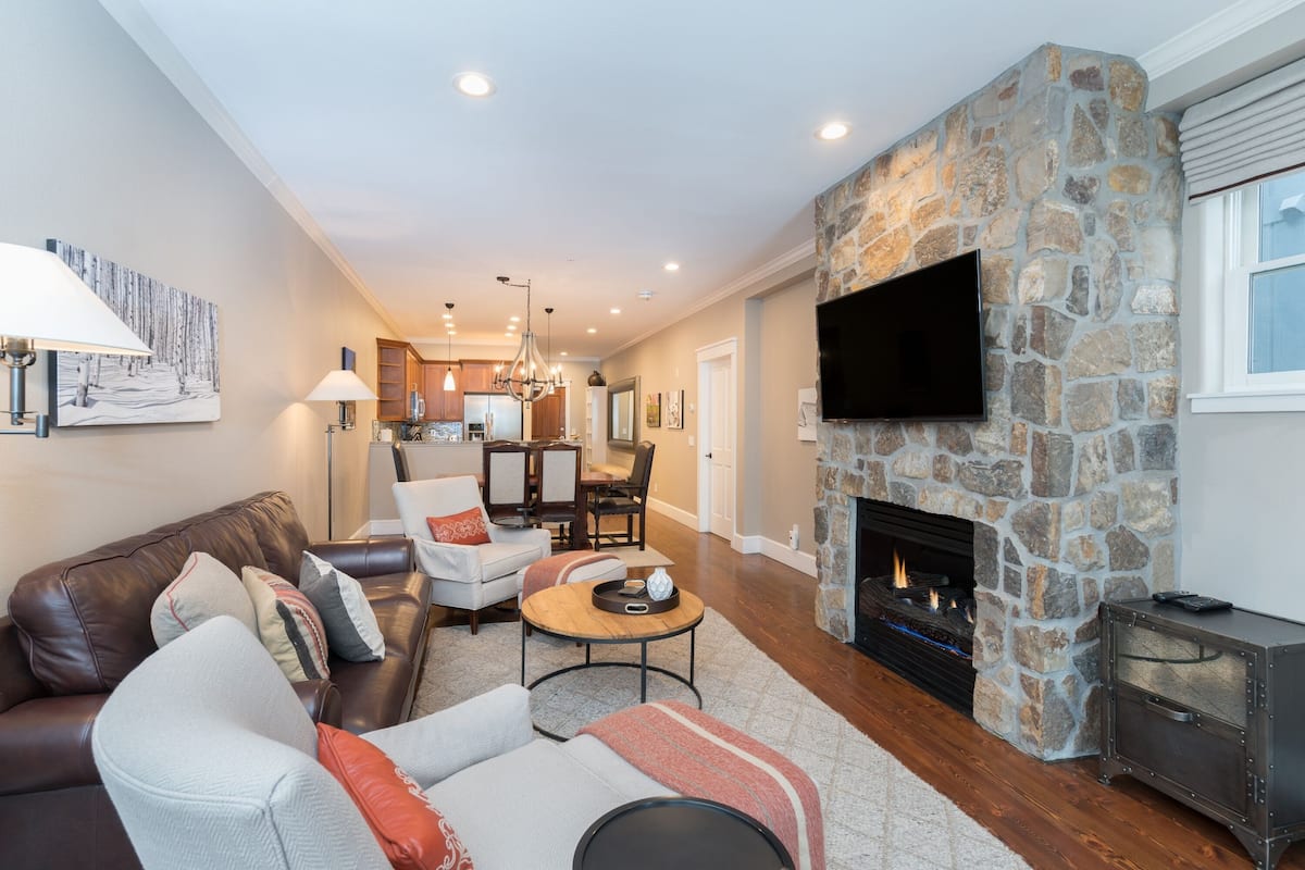 A modern living room with a stone fireplace, leather sofa, and a flat-screen TV mounted on the wall.