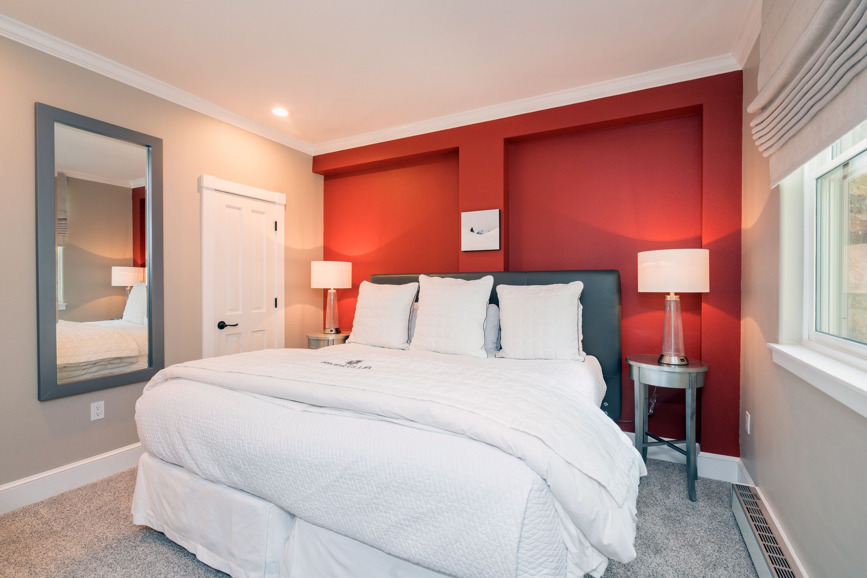 Un dormitorio con una cama grande, dos mesitas de noche con lámparas, un espejo y una pared de acento roja.