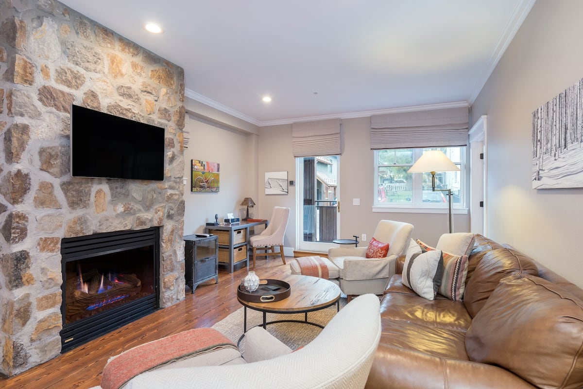 A living room with a stone fireplace, leather sofa, and a flat-screen TV mounted on the wall.