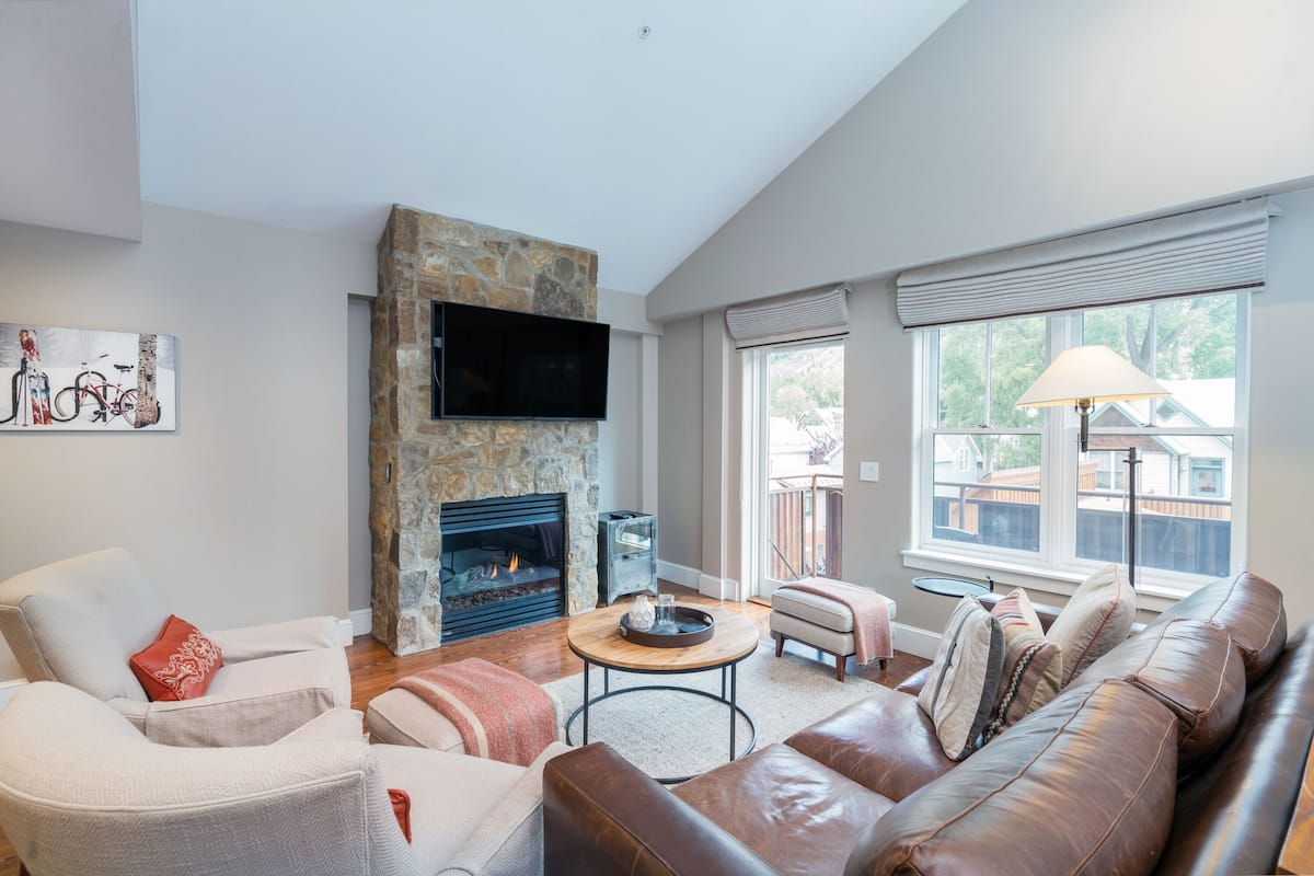 A living room with a stone fireplace, leather sofa, and a flat-screen TV mounted on the wall.