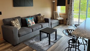 A modern living room with a grey sofa, a coffee table, a dining table with chairs, a large window with curtains, and a wall-mounted abstract art piece. - Connect with nature - Tranquil Downeast Escape, Near Acadia & Sunrise Trail (Eastbrook)