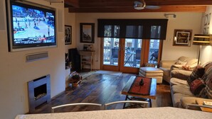 A living room with a TV displaying a basketball game, a fireplace, a dining area, and a view through the glass doors. - Modern Contemporary Condo-Tahoe Lodge Atmosphere in Incline Village, NV. (Incline Village)