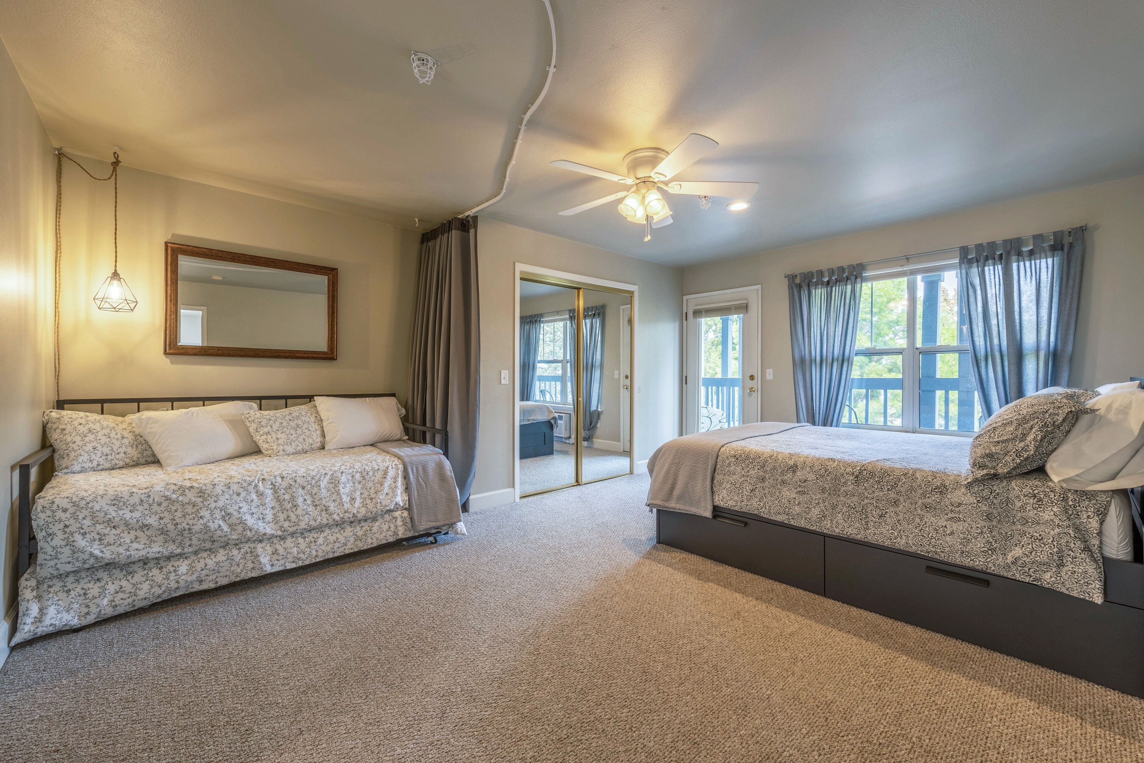 A bedroom with two beds, a ceiling fan, a large mirror, and a view of the outside through a window.