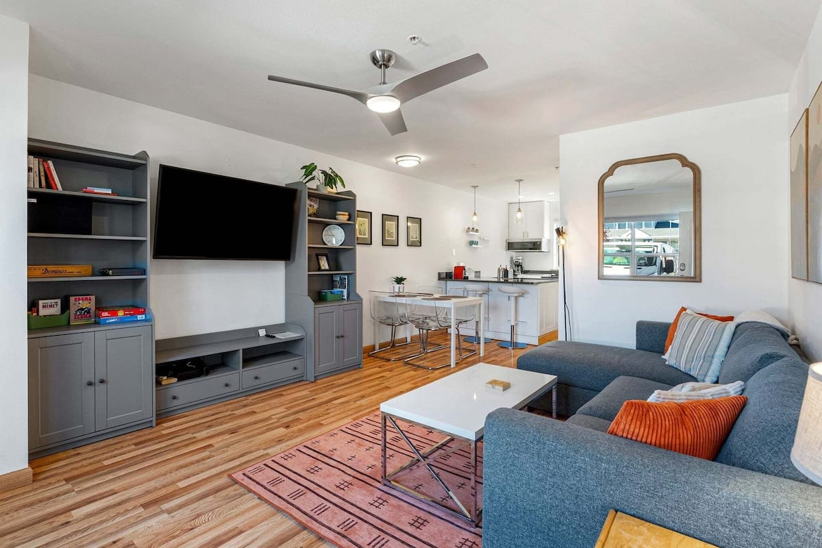 A modern living room with a grey sofa, a white coffee table, a flat-screen TV, a ceiling fan, and a kitchen area in the background.