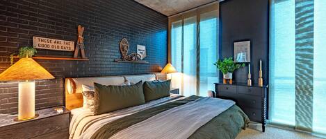 A modern bedroom with a brick wall, a large bed, two bedside tables with lamps, a dresser, and a large window with curtains.