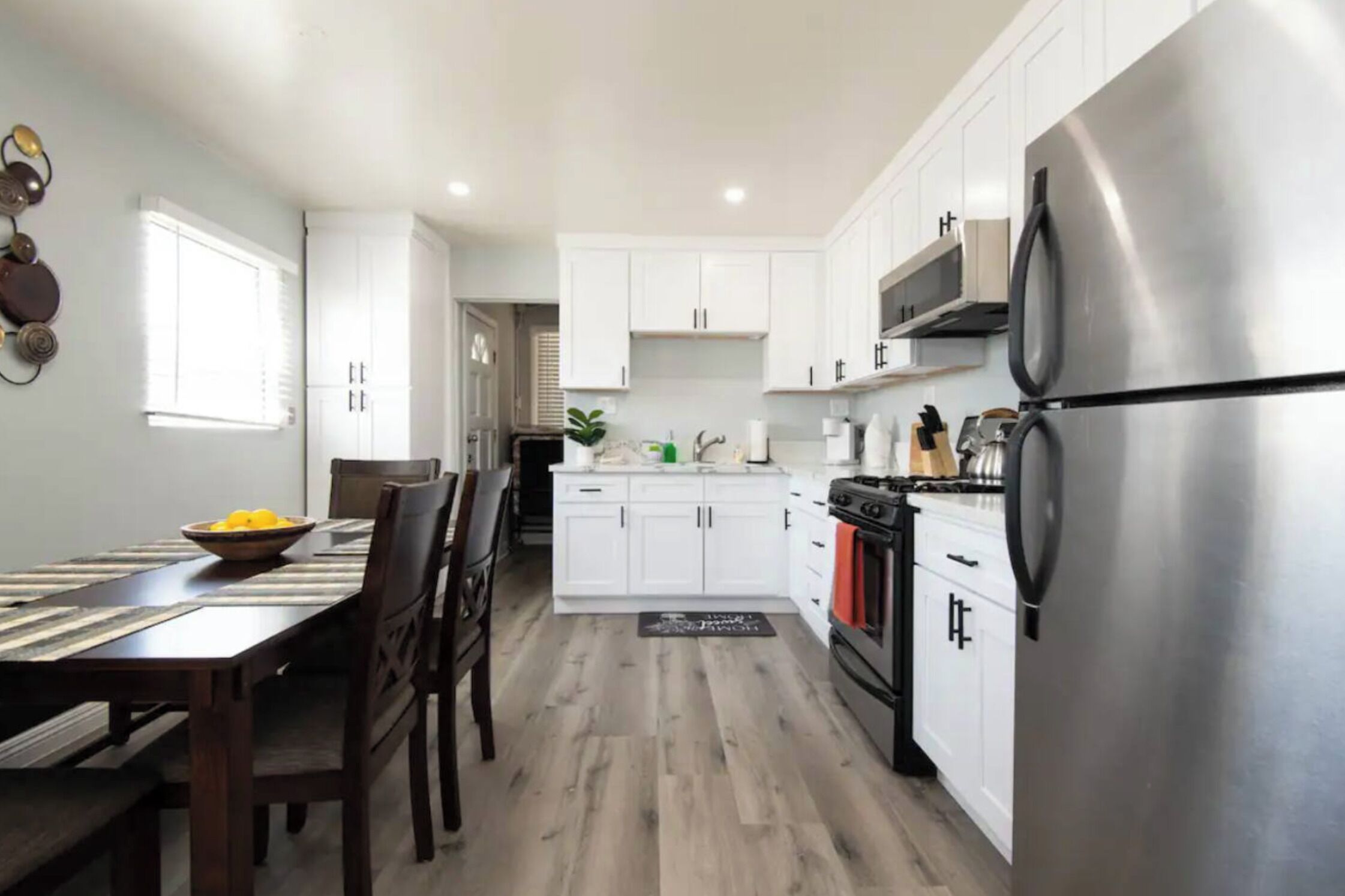 Una cocina moderna con armarios blancos, un refrigerador de acero inoxidable y una zona de comedor con una mesa y sillas de madera.