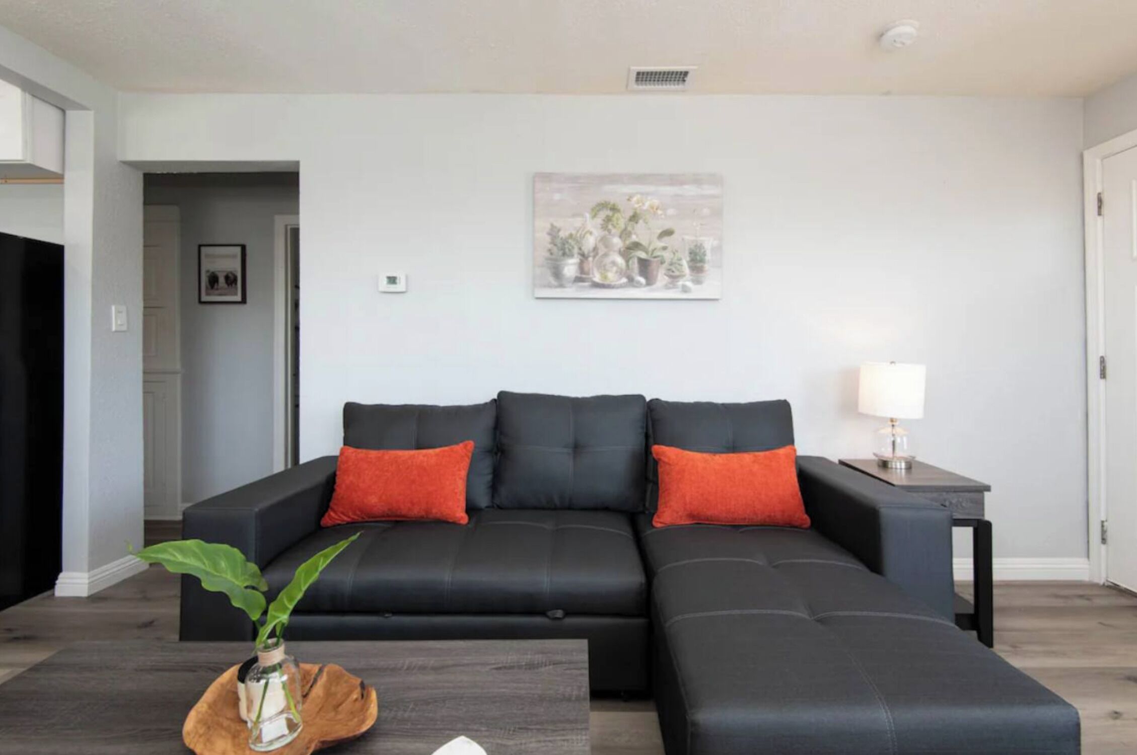 A modern living room with a black sectional sofa, two red cushions, a small table with a lamp, and a plant on a wooden coffee table.
