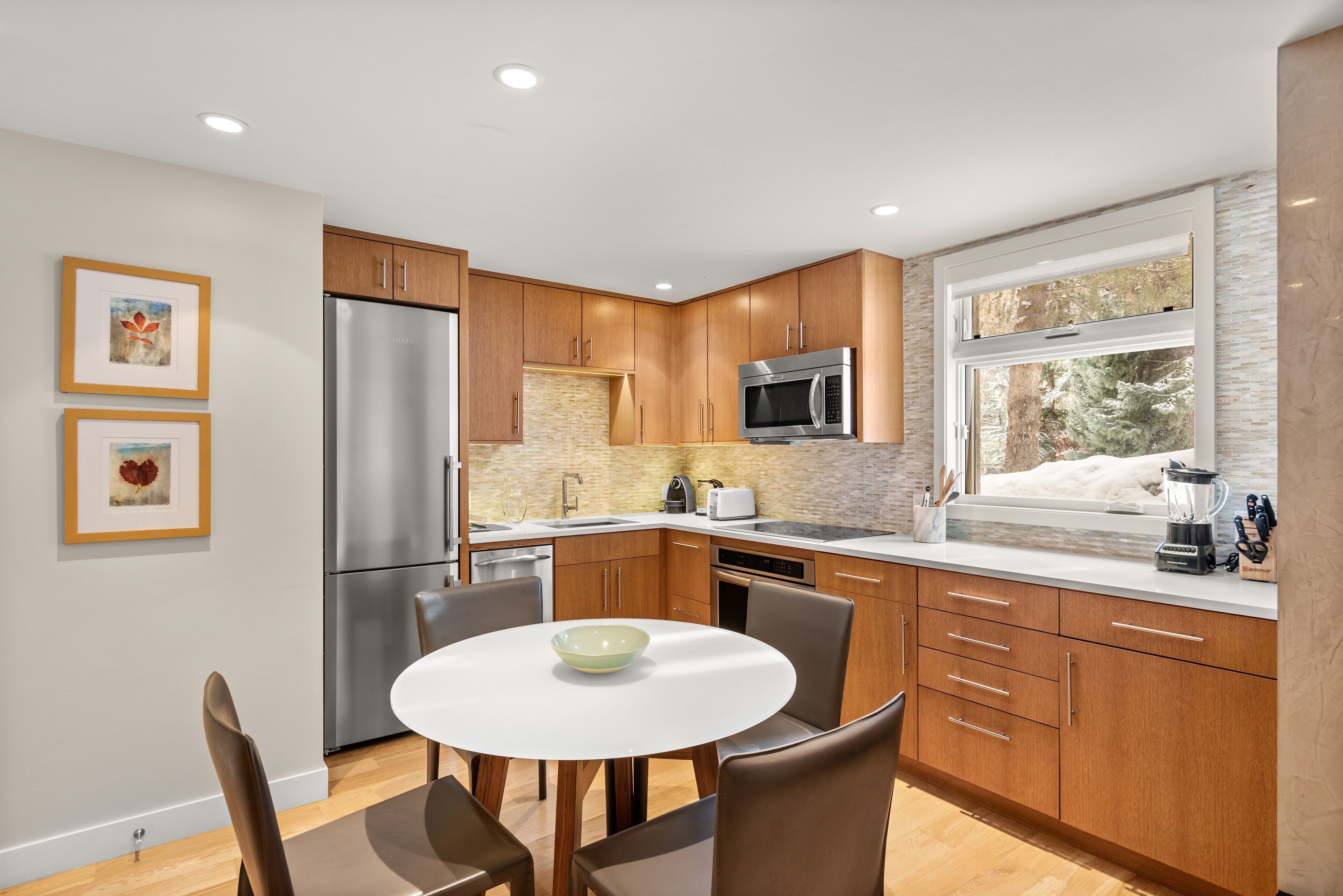 Una cocina moderna con armarios de madera, electrodomésticos de acero inoxidable, una mesa de comedor blanca y un ventanal con vistas a árboles.