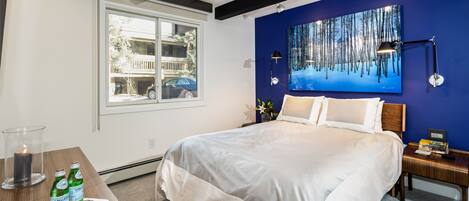 Une chambre avec un grand lit, une fenêtre donnant sur un balcon et une voiture garée, un tableau accroché au mur, et une table de chevet avec une lampe et un livre.