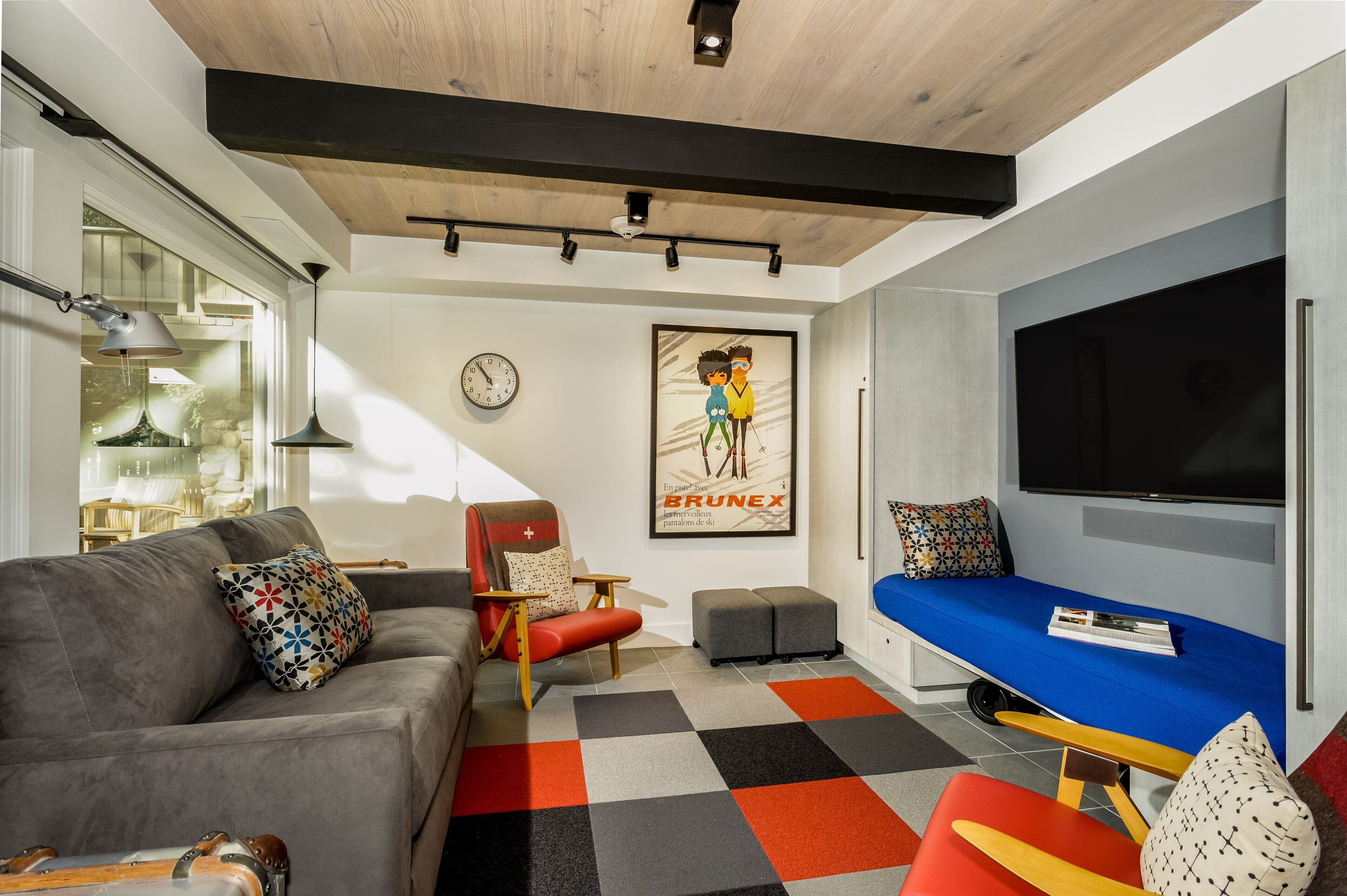 A modern living room with a grey sofa, red armchair, and blue sofa bed. There is a large flat-screen TV mounted on the wall, a clock, and a poster. The room has a wooden ceiling and a patterned rug.