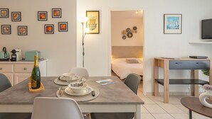 Quarto moderno e bem iluminado com uma mesa de jantar posta para dois, uma cama com cabeceira, uma escrivaninha com computador e diversas obras de arte emolduradas nas paredes.
