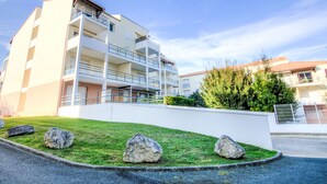 Um edifício residencial com vários andares, varandas, um gramado bem cuidado e pedras grandes em primeiro plano.