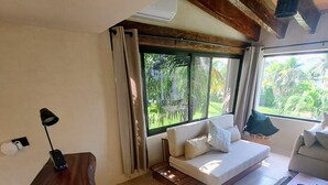 A modern living room with a curved wooden desk, a sofa with cushions, a small wooden table, and a view of greenery through large windows. - Cancún Energy, Tulum Spirit  Luxe & Private Stay (Cancún)