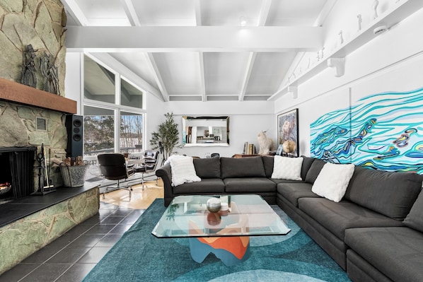 A modern living room with a stone fireplace, a glass coffee table, a dark sofa, and a large abstract painting on the wall.