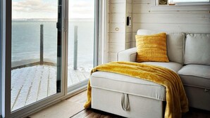 Una habitación con puerta de vidrio corredera que da a una terraza, un sofá con una manta color mostaza y una ventana pequeña con un marco de fotos.