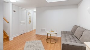 A modern living room with a grey sofa, a small round table with a book, and a patterned rug. A staircase with wooden steps leads to another room. - Newly Built Basement Suite – Comfort & Convenience (Markham)