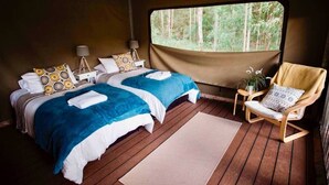 Ein gemütliches Schlafzimmer mit zwei Betten, Holzboden, Fenster mit Blick auf den Wald, einem Sessel und einem kleinen Tisch mit einer Lampe.