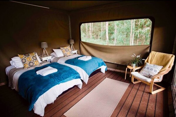 A cozy bedroom with two beds, a wooden floor, a window with a forest view, a chair, and a small table with a lamp.