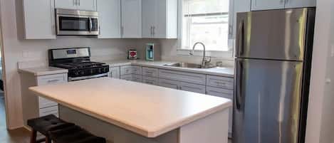 A modern kitchen with white cabinetry, a stainless steel refrigerator, a microwave, and a stove. There are three bar stools at the island.