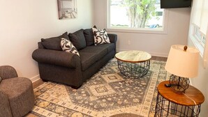 A living room with a dark grey sofa, patterned cushions, a round coffee table, a small side table with a lamp, a patterned rug, and a TV mounted on the wall. - LakePointe Shores # 7 Welcoming 3-bedroom cottage in Madison on the Lake (Madison)