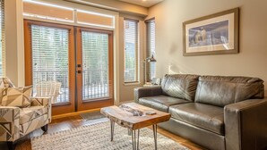 A living room with a leather sofa, a patterned armchair, a coffee table, and a large window with blinds. - Sun Peaks Resort BC - Stones Throw Vacation Rentals (Sun Peaks)