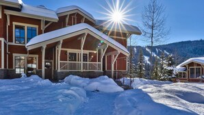 A large, two-story lodge with a covered porch and a ski slope in the background. - Sun Peaks Resort BC - Stones Throw Vacation Rentals (Sun Peaks)
