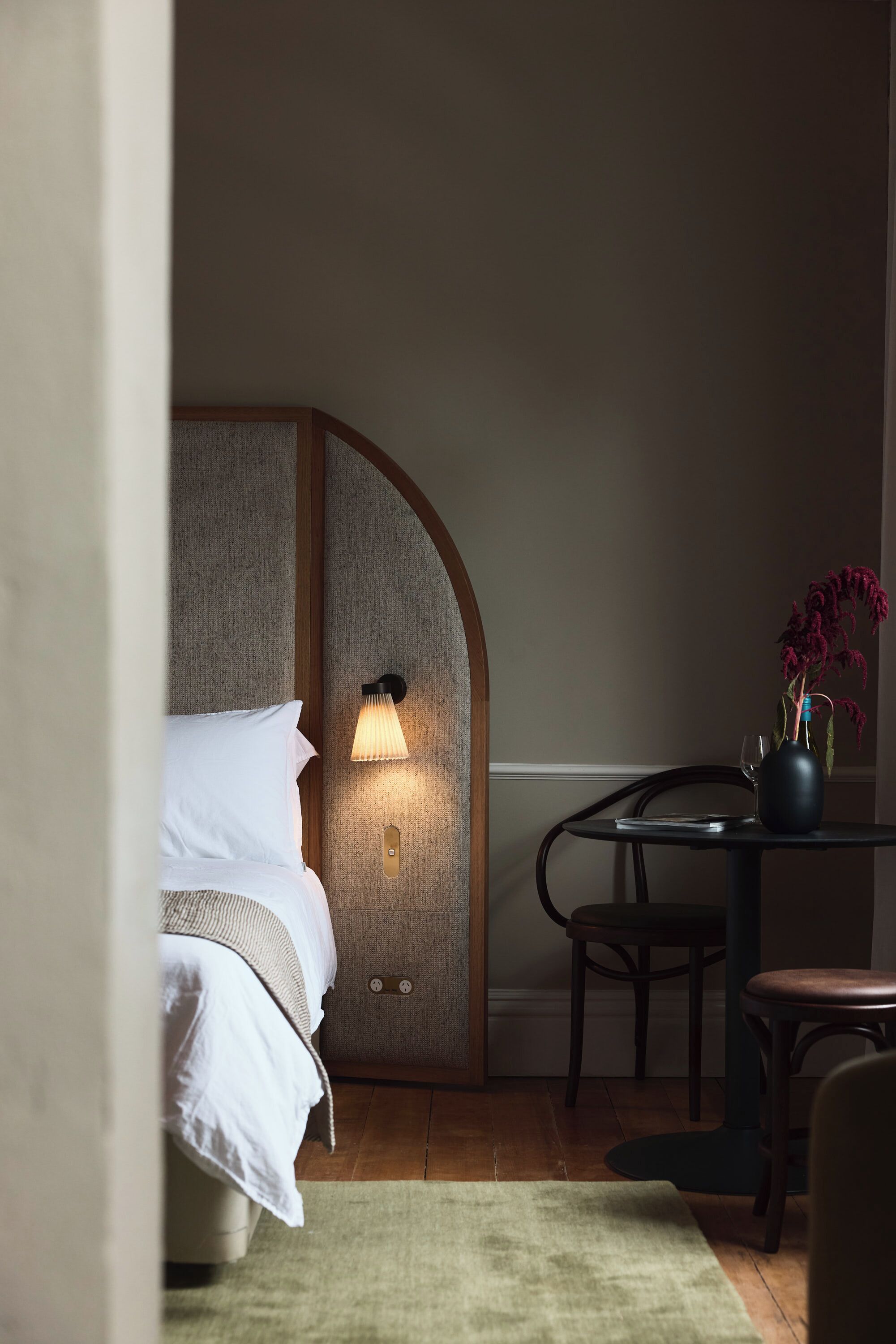 A hotel room with a bed, a nightstand, a table with a vase, and a lamp.