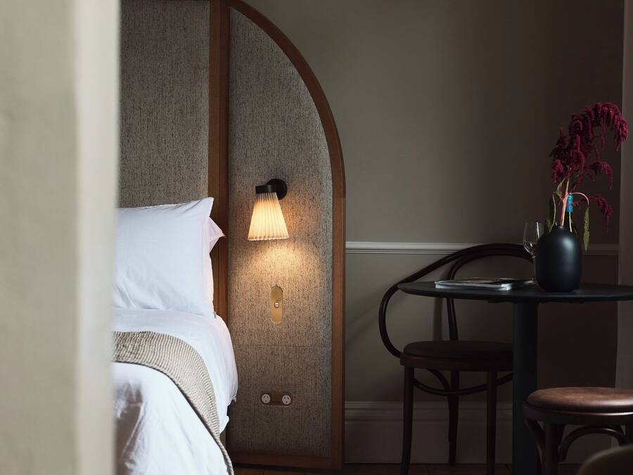A hotel room with a bed, a nightstand, a table with a vase, and a lamp.