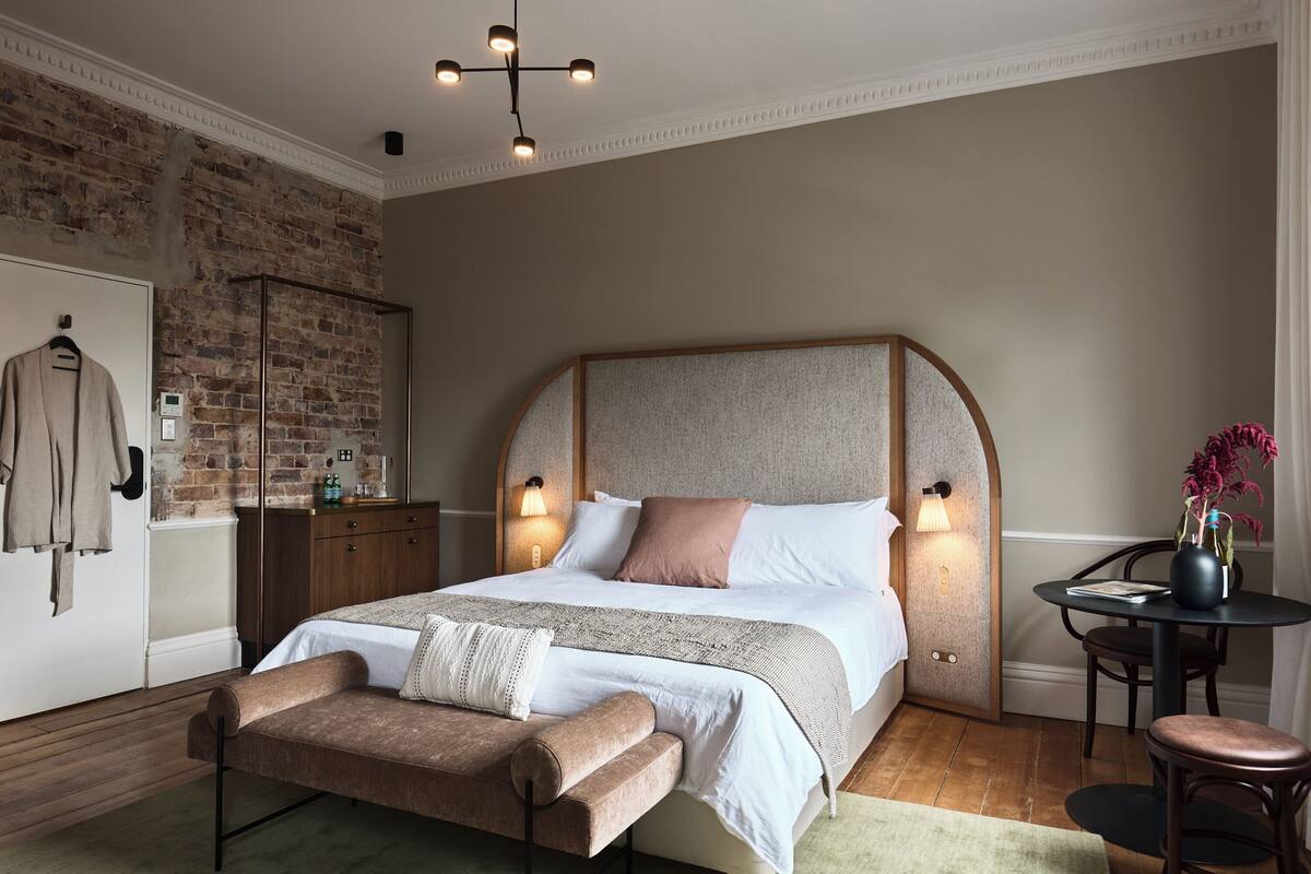 A bedroom with a large bed, a wooden bench, a small table with a vase, and a brick wall.