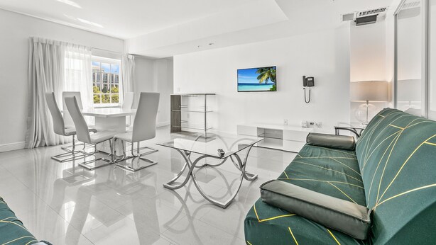 A modern living room with a glass coffee table, a sofa with geometric patterns, a dining area with a white table and chairs, and a large window with a view of a beach. - NEW LISTING! 4 Bedroom Combined Suites (Miami Beach)