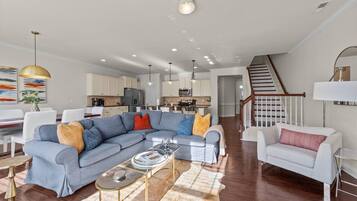 Una sala de estar moderna con un sofá seccional gris, un sillón blanco y una mesa de centro. Se observa una escalera que lleva al piso superior, y un área de cocina al fondo.
