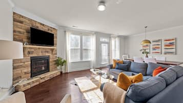A modern living room with a stone fireplace, a flat-screen TV, a grey sofa with colorful cushions, and a dining area with a table and chairs.