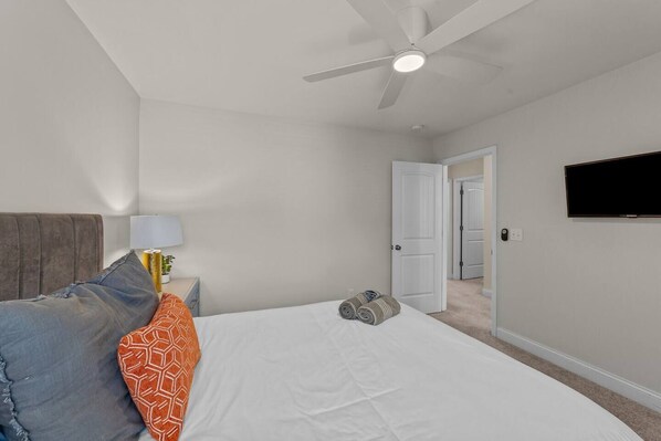 A bedroom with a bed, pillows, a nightstand, a lamp, a ceiling fan, a TV, and a door leading to another room.
