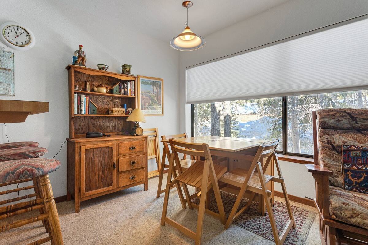 Een gezellige kamer met een houten eettafel, stoelen, een boekenkast en een groot raam met uitzicht op bomen.