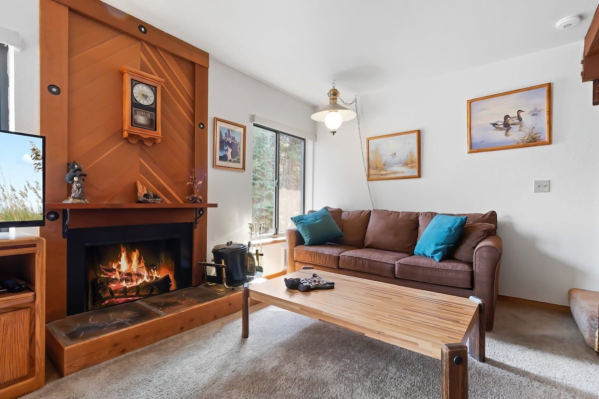 Een woonkamer met een open haard, een bruine bank met blauwe kussens, een houten salontafel en lijstwerken aan de wanden.