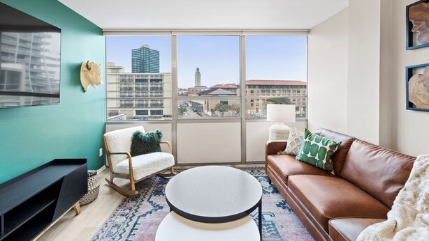 Una sala de estar moderna con un ventanal grande, un sofá de cuero marrón, un sillón blanco, una mesa de centro redonda y un mueble negro para televisor.