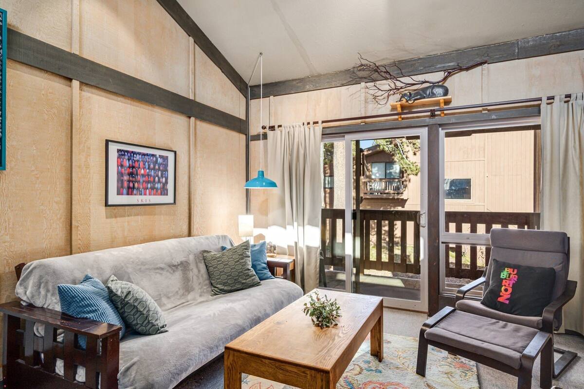 Ein gemütlicher Wohnraum mit einer Couch, einem Holztisch und einem Sessel. An der Wand hängt ein gerahmtes Bild und es gibt ein Fenster mit Vorhängen.