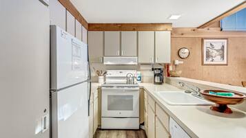 A compact kitchen with white appliances, wooden beams, a sink, and a clock on the wall.