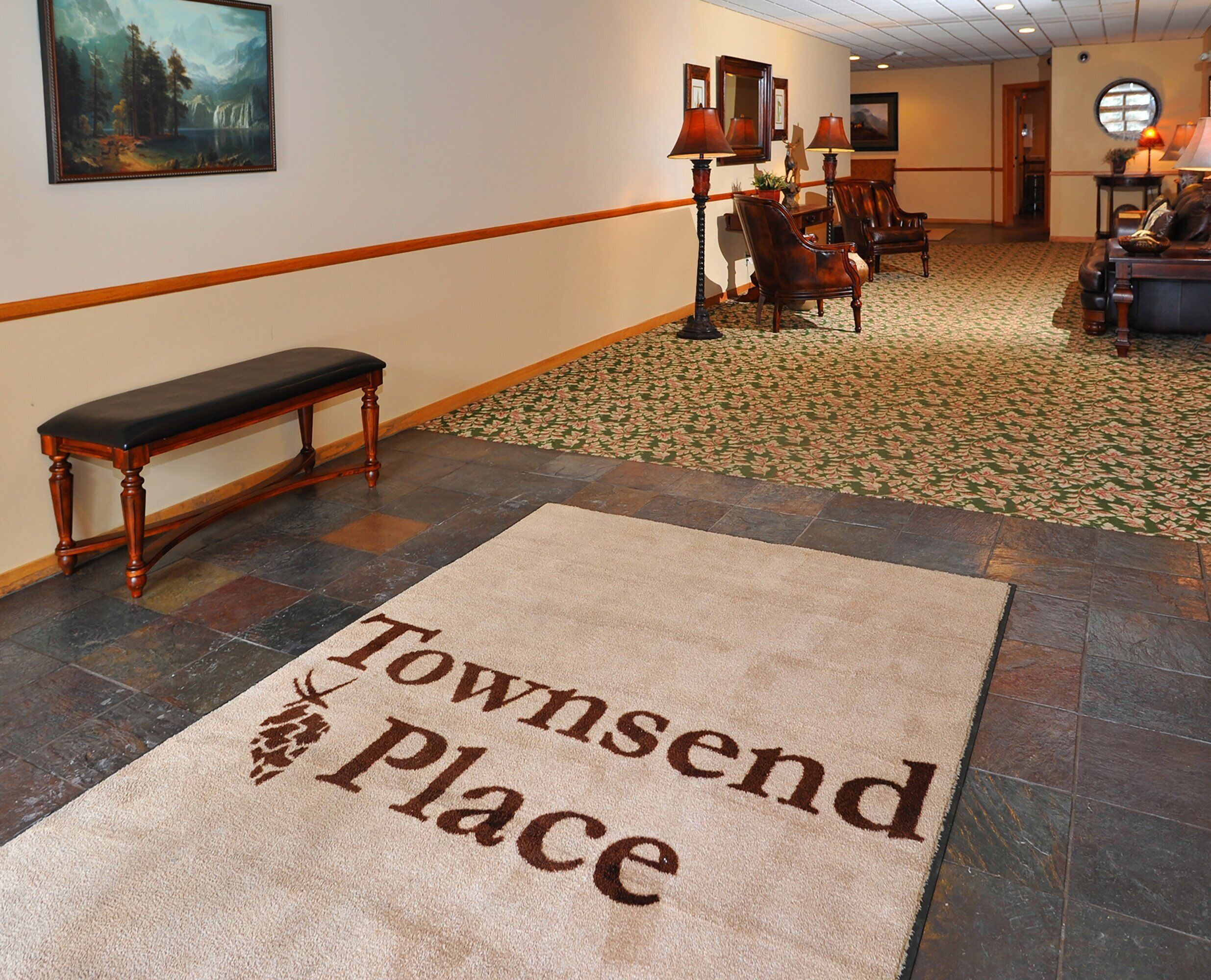 Um saguão de hotel com um banco, cadeiras, um tapete com o texto 'Townsend Place' e uma pintura na parede.