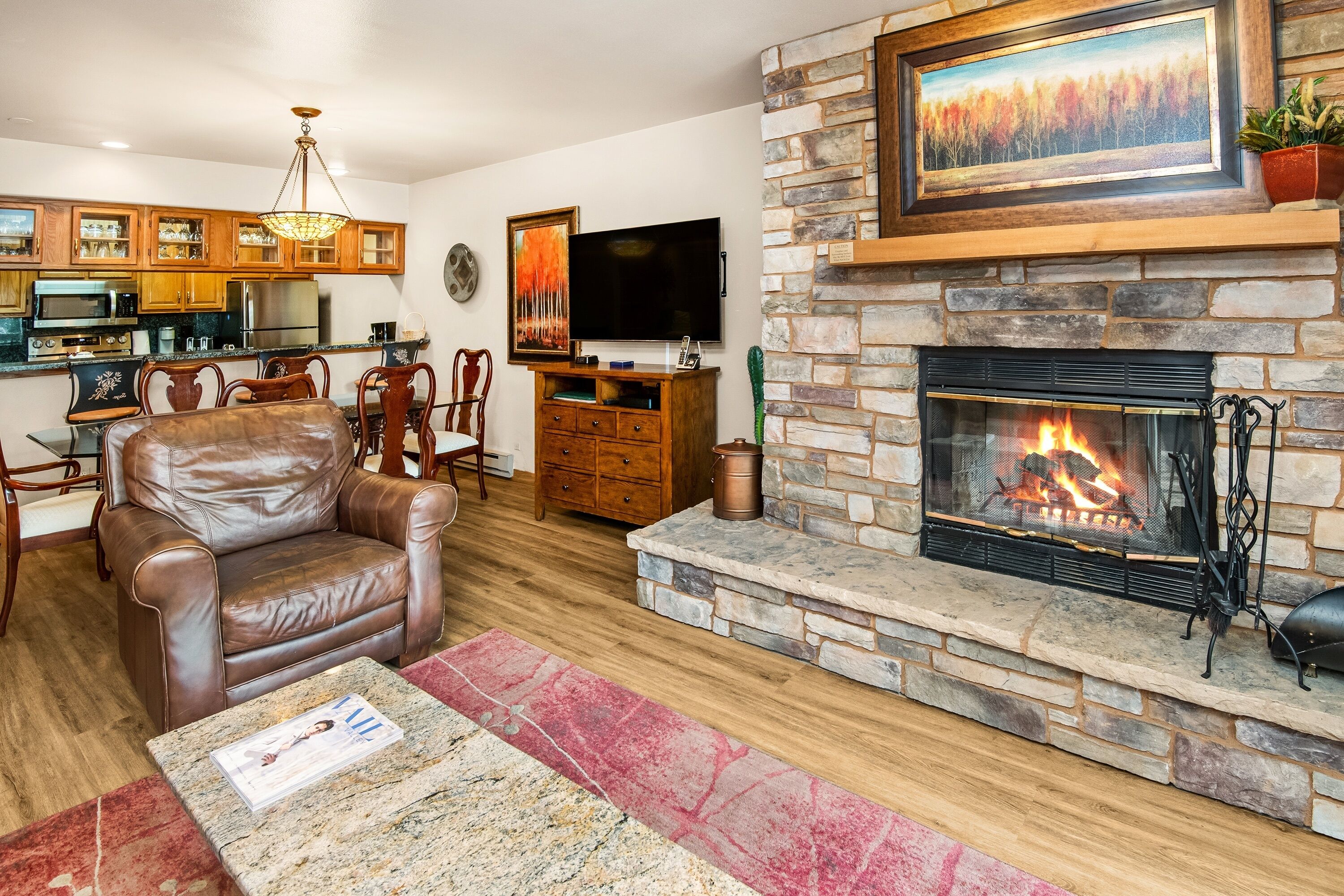 A living room with a stone fireplace, leather armchair, dining area, kitchen, and a flat-screen TV.