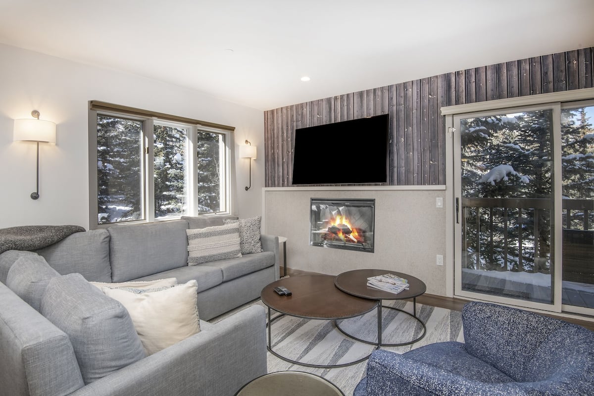 A modern living room with a fireplace, grey sofas, a coffee table, and large windows with a snowy view.