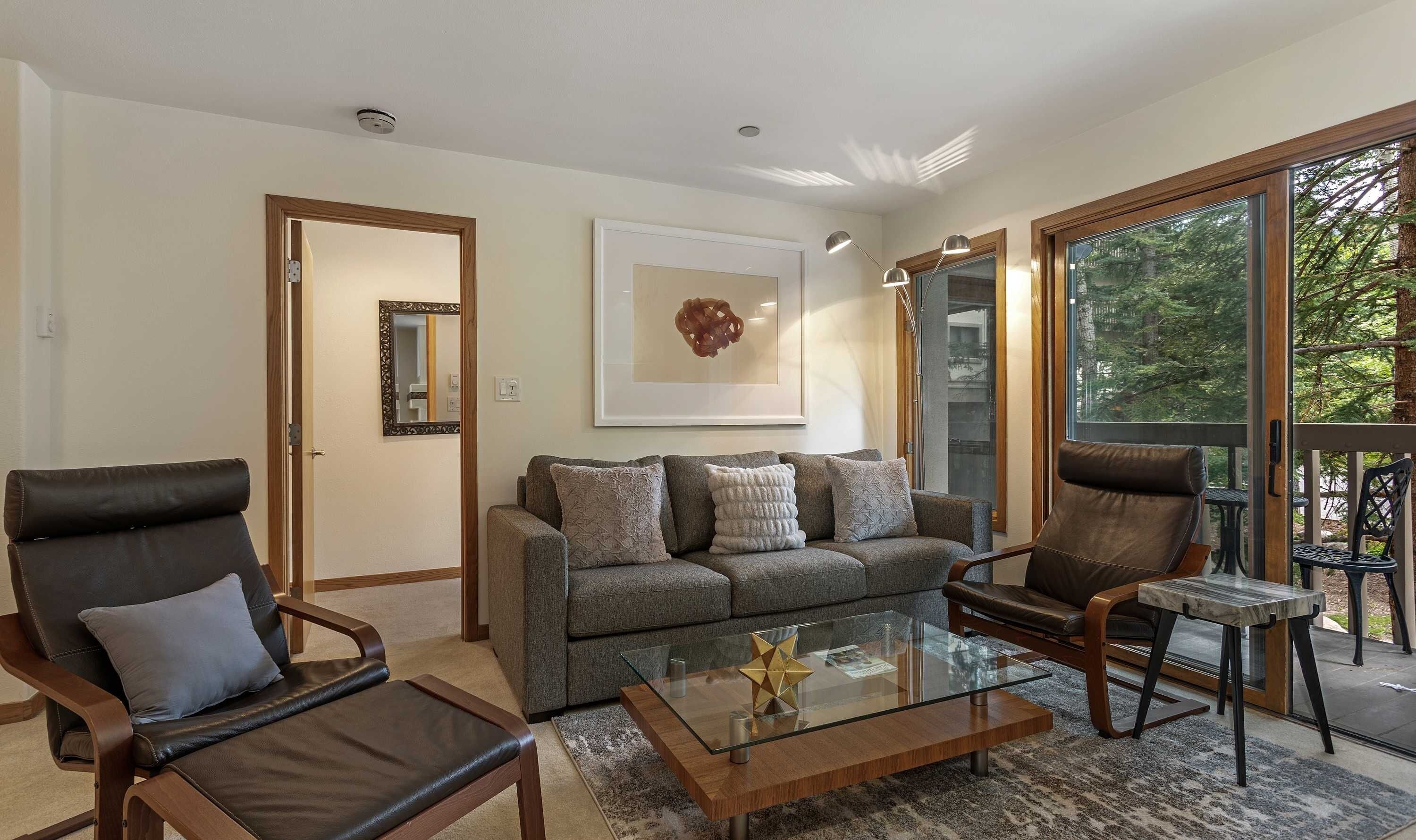 A modern living room with a sofa, two leather chairs, a glass coffee table, and a large window with a view of trees.