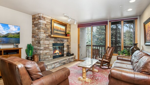 A living room with leather sofas, a stone fireplace, a flat-screen TV, and a large window with a view of trees. - Beaver Creek Resort CO - Townsend Place Vacation Rentals (Avon)