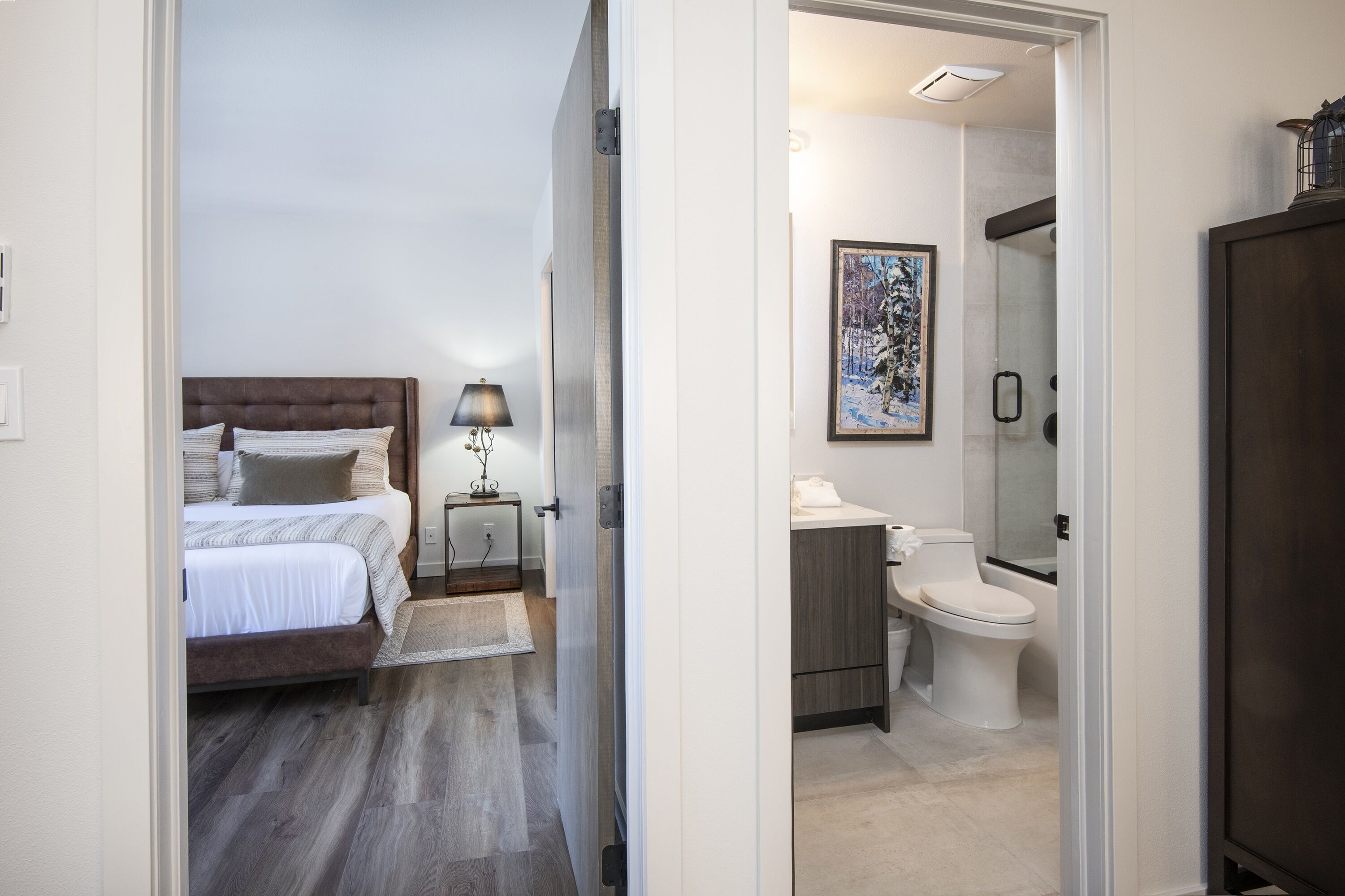A bedroom with a bed, bedside table, and lamp. The room has a wooden floor and a door leading to a bathroom with a toilet and sink.