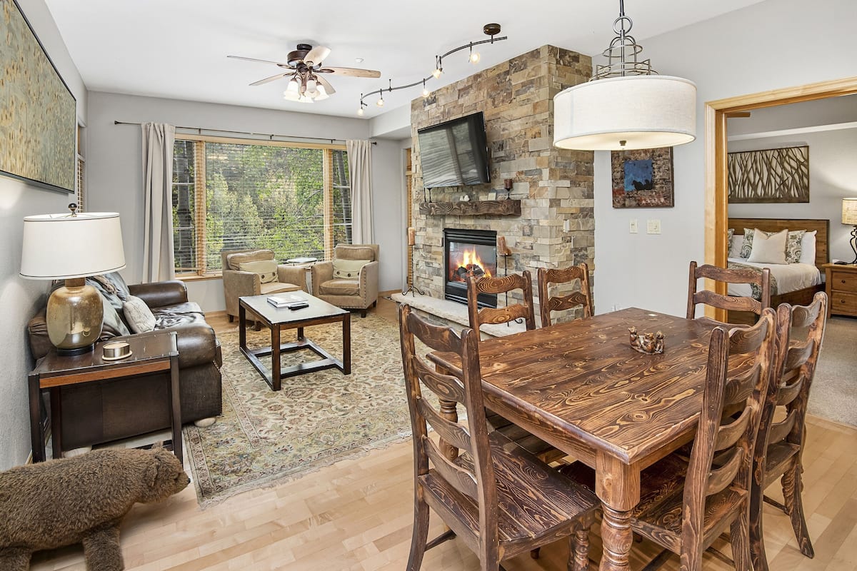 A spacious living room with a fireplace, a dining area, and a large window offering a view of trees.